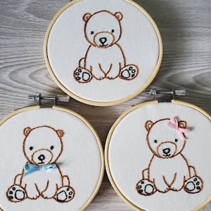 May include: Three embroidered hoop art pieces featuring brown teddy bears. The middle bear is wearing a blue bow tie, the right bear is wearing a pink bow.