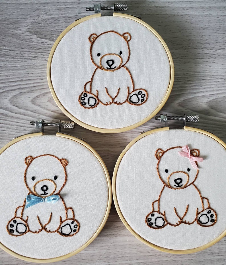 May include: Three embroidered hoop art pieces featuring brown teddy bears. The middle bear is wearing a blue bow tie, the right bear is wearing a pink bow.