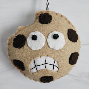 May include: A brown felt cookie with a bite taken out of it. The cookie has black chocolate chips and a white felt face with black button eyes and a stitched mouth.