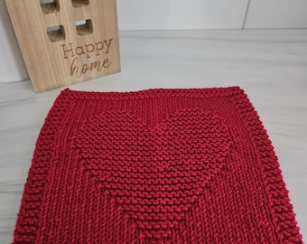 May include: A red knitted square with a heart design in the center. The heart is formed by the pattern of the knit stitches. The square is surrounded by a border of the same red yarn. A wooden house with the words "Happy home" is in the background.