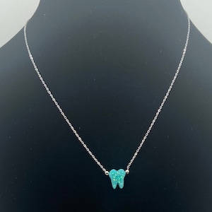 May include: A silver chain necklace with a small, turquoise tooth pendant. The tooth pendant has a textured surface and is centered on the chain. The necklace is displayed against a black background.