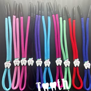 May include: A collection of colorful eyeglass holders. Each holder features a cord in a different color, including blue, pink, black, purple, green, and red. Each cord has a white tooth-shaped charm with a toothpaste design. The text "Tooth Fairy Store" is visible.