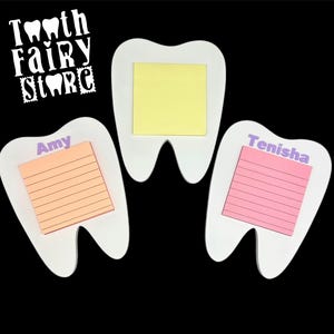 May include: Three white tooth-shaped note holders with sticky notes. The top note holder has a yellow sticky note with no text. The left note holder has an orange sticky note with the name "Amy" written in purple. The right note holder has a pink sticky note with the name "Tenisha" written in purple.