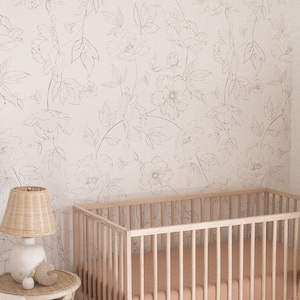 May include: A white crib with a light pink mattress, a white wicker bassinet, a white armchair with a cream pillow, a woven ottoman, and a white lamp with a wicker shade. The room has white walls with a floral wallpaper pattern and a white ceiling fan with a woven shade.