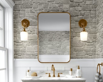 May include: A bathroom scene featuring a gray vanity with gold hardware, a rectangular gold-framed mirror, and two sconces. The walls are adorned with a stone-like wallpaper. A striped towel hangs on a gold ring.
