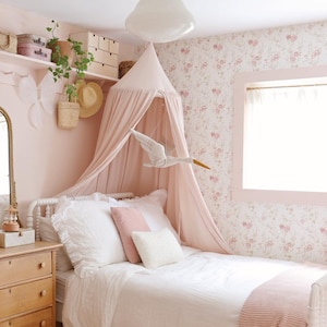 A cozy, vintage-style girl bedroom with floral wallpaper, a white bed with pink canopy, wooden dresser, and decorative items on shelves. The room has a warm, inviting feel with soft pink and white tones.
