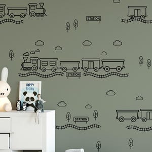 May include: Black line art train wall decals with clouds, trees, and station signs on a green wall. A white cabinet holds a bunny lamp, framed panda art, and stuffed animals. Baskets and toys are on the floor.