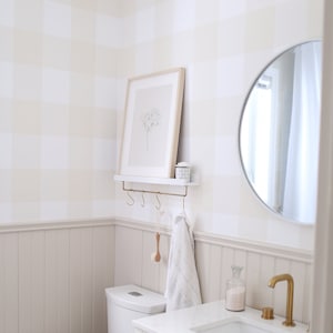 Peut inclure: Une salle de bain blanche avec des toilettes blanches, une vasque blanche avec un comptoir blanc et un robinet doré, un miroir rond et une estampe encadrée sur le mur. Les murs sont recouverts d'un papier peint vichy beige et blanc.