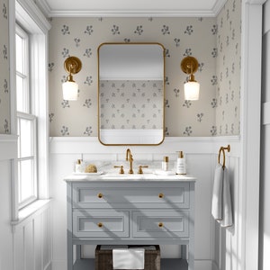May include: A grey bathroom vanity with three drawers and a lower shelf. The vanity has gold hardware and a white countertop. A gold framed mirror hangs above the vanity. The walls are covered in a beige wallpaper with a blue floral pattern.