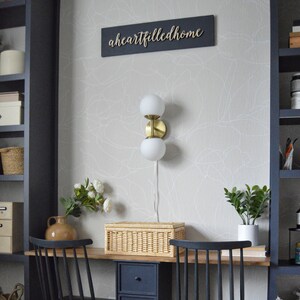 May include: A home office desk with a black and wood finish. The desk has a wicker storage box, a gold wall sconce with two white globe lights, and a potted plant. The wall behind the desk has a white and gray abstract wallpaper with the text "aheartfilledhome" on a black sign.