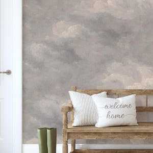 Könnte beinhalten: Eine Holzbank mit einem gestreiften Kissen und einem weißen Kissen mit der Aufschrift "welcome home" in Schreibschrift. Neben der Bank stehen zwei olivgrüne Gummistiefel. Die Wand dahinter zeigt ein Wandbild mit bewölktem Himmel.