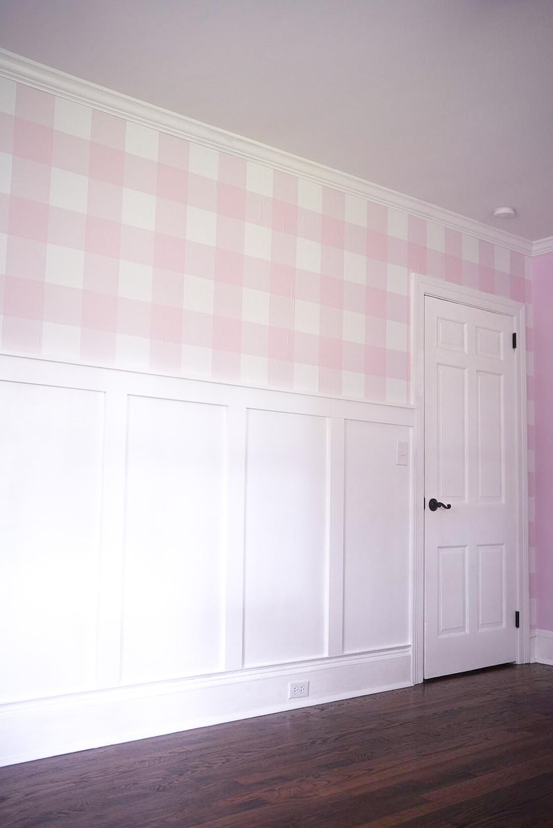 May include: A wall featuring a light pink and white gingham pattern on the upper section, with white wainscoting on the lower portion. A white six-panel door with a black handle is integrated into the wall, set against dark wood flooring.