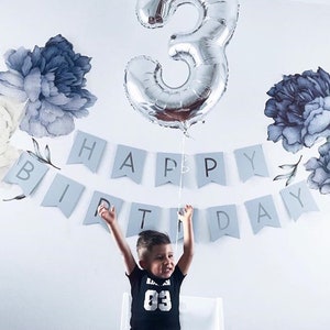 May include: A child wearing a black shirt with the number "03" on it, is standing in front of a white wall with a blue and white banner that says "Happy Birthday". A silver number "3" balloon is floating above the child's head.