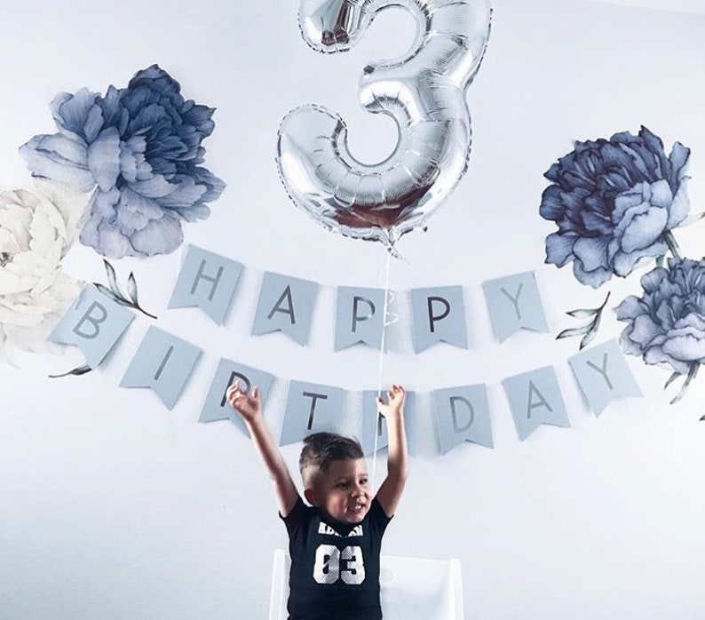May include: A child wearing a black shirt with the number "03" on it, is standing in front of a white wall with a blue and white banner that says "Happy Birthday". A silver number "3" balloon is floating above the child's head.