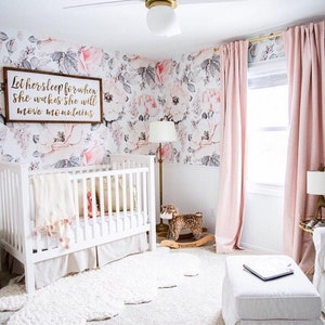 May include: A white crib with a white and pink blanket and a white rug in a nursery with a floral wallpaper, a pink rocking horse, and a pink sign that says "Let her sleep for when she wakes she will move mountains."