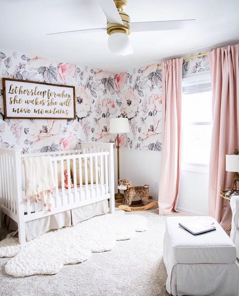 May include: A white crib with a white and pink blanket and a white rug in a nursery with a floral wallpaper, a pink rocking horse, and a pink sign that says "Let her sleep for when she wakes she will move mountains."