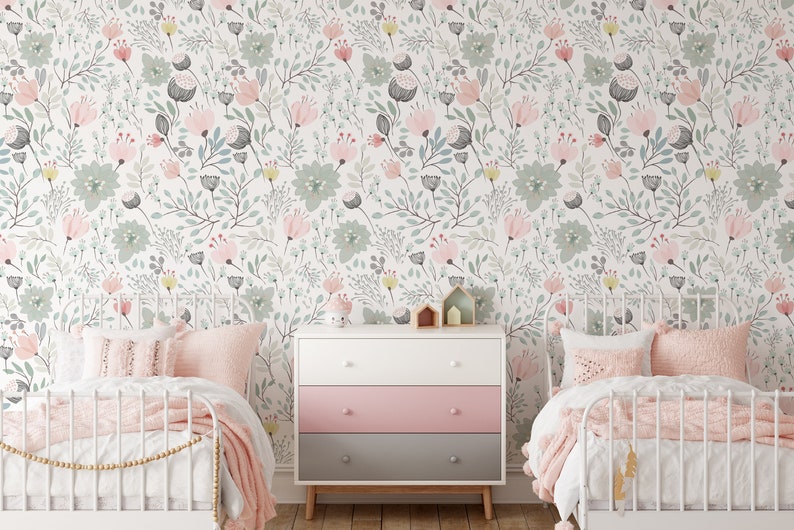 May include: Two white twin beds with pink bedding and pillows in a room with a white dresser with pink and gray drawers. The walls are covered in a floral wallpaper with pink, green, and yellow flowers.