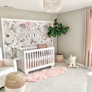 May include: A white crib with a floral wallpaper backdrop. The wallpaper features pink and gray flowers. The name "Lilly" is spelled out in white letters above the crib. A pink and white rug is on the floor in front of the crib.