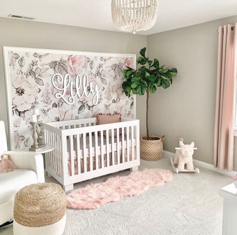 May include: A white crib with a floral wallpaper backdrop. The wallpaper features pink and gray flowers. The name "Lilly" is spelled out in white letters above the crib. A pink and white rug is on the floor in front of the crib.