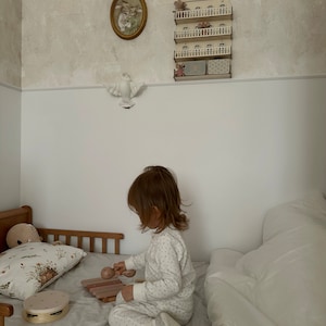以下が含まれることがあります： 小さな子供がベッドに座って、2本のバチで木製のシロフォンを演奏しています。シロフォンはピンクと茶色です。子供は白とグレーの模様の入ったパジャマの上下を着ています。ベッドには白い羽毛布団と花柄の枕があります。