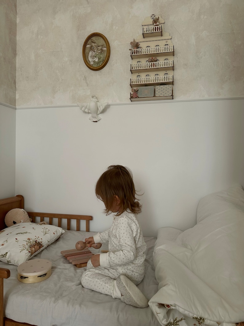 以下が含まれることがあります： 小さな子供がベッドに座って、2本のバチで木製のシロフォンを演奏しています。シロフォンはピンクと茶色です。子供は白とグレーの模様の入ったパジャマの上下を着ています。ベッドには白い羽毛布団と花柄の枕があります。