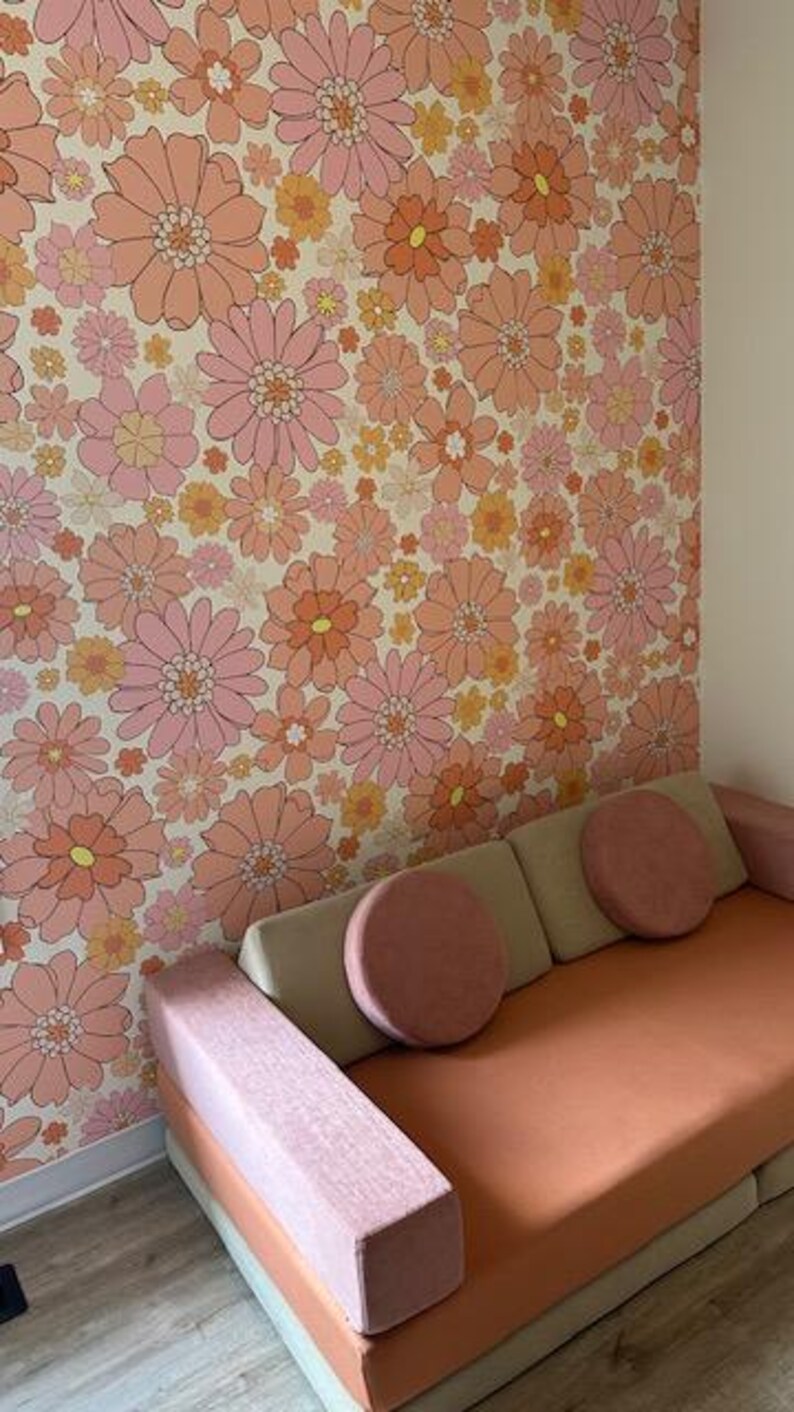 May include: A pink and orange floral patterned wallpaper with a light pink and beige couch with two pink pillows. The couch has a light pink armrest.