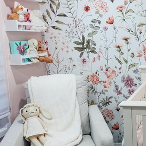 May include: A white armchair with a plush white blanket and a stuffed bunny doll. The armchair is in front of a wall with a floral wallpaper featuring pink, red, and green flowers.