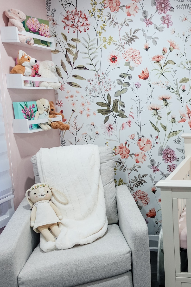 May include: A white armchair with a plush white blanket and a stuffed bunny doll. The armchair is in front of a wall with a floral wallpaper featuring pink, red, and green flowers.