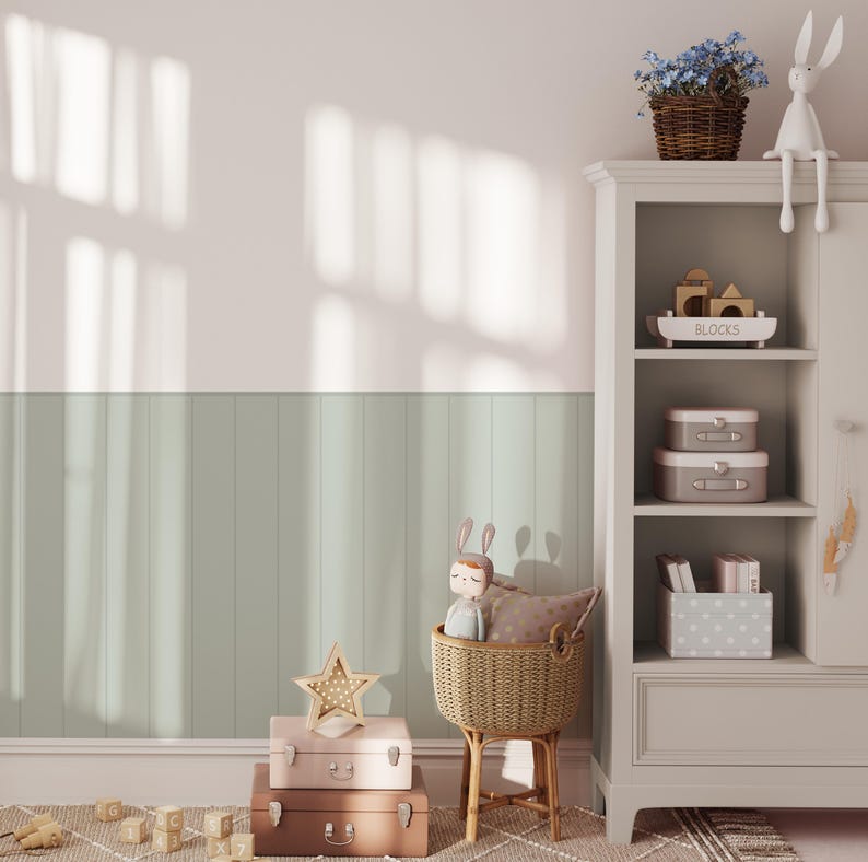 May include: A light gray wooden shelf unit with three shelves. The top shelf has a wooden box with the word "BLOCKS" on it. The middle shelf has two stacked boxes, one pink and one gray. The bottom shelf has a stack of books and a gray box with white polka dots. A wicker basket with a stuffed toy rabbit is on the floor in front of the shelf. There are three stacked suitcases on the floor in front of the basket.