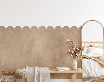 May include: A light brown textured wall with a scalloped border. A wooden bench with a white throw blanket sits in front of the wall. A vase with dried flowers sits on the bench. A large mirror with a gold frame is in the background.