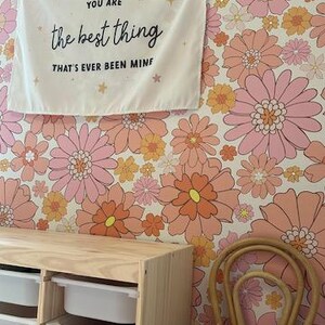 May include: A white canvas wall tapestry with the text "You are the best thing that's ever been mine" in black lettering. The tapestry is hanging on a wall with a pink and orange floral wallpaper. A wooden storage unit with white drawers and a wicker chair are in the foreground.