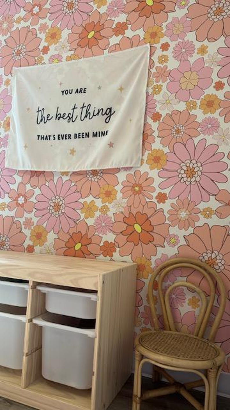 May include: A white canvas wall tapestry with the text "You are the best thing that's ever been mine" in black lettering. The tapestry is hanging on a wall with a pink and orange floral wallpaper. A wooden storage unit with white drawers and a wicker chair are in the foreground.