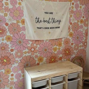 May include: A wooden storage unit with three white bins. The unit is in front of a wall with a floral wallpaper featuring pink and orange flowers. A sign above the unit reads "You are the best thing that's ever been mine."