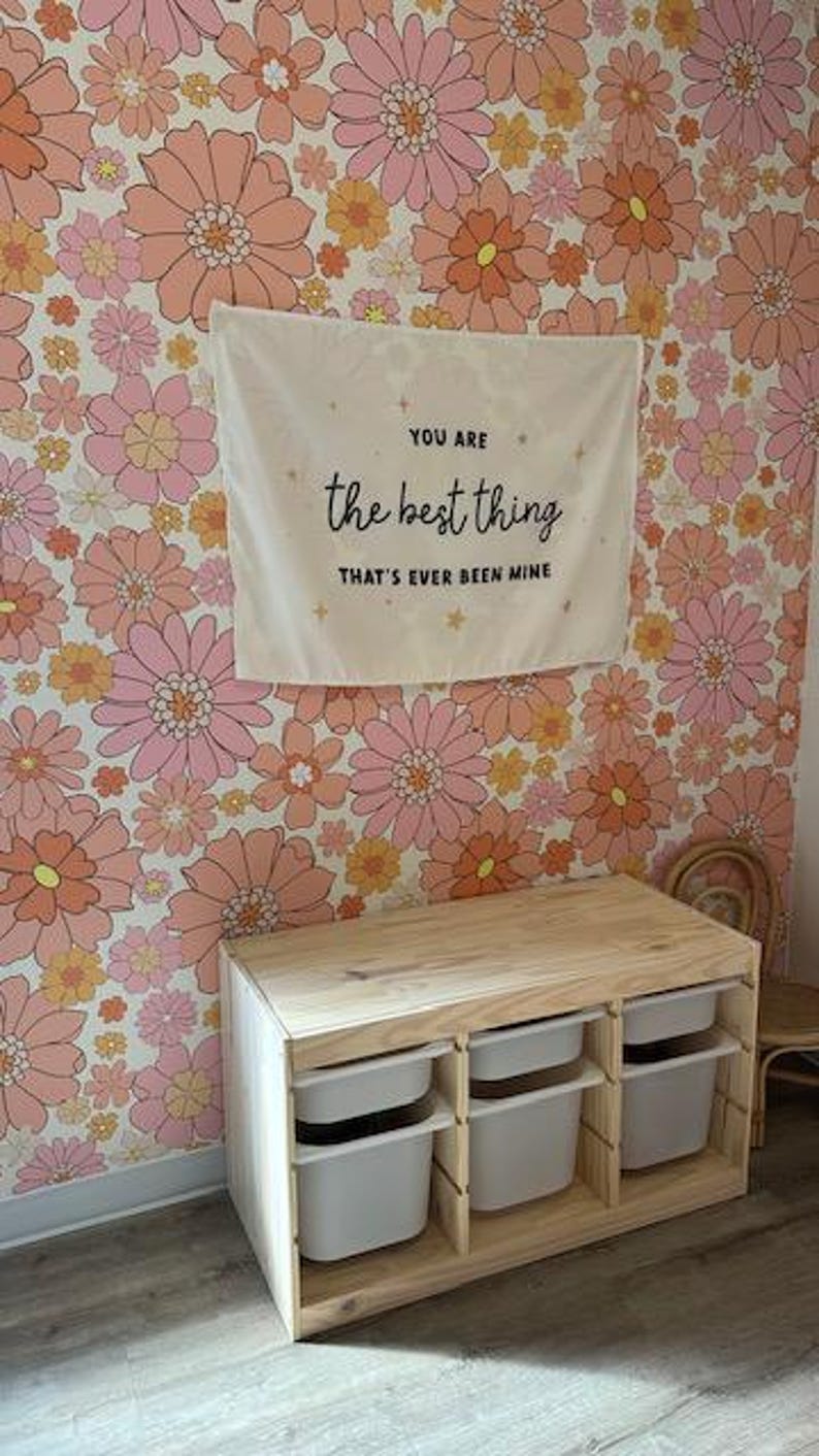 May include: A wooden storage unit with three white bins. The unit is in front of a wall with a floral wallpaper featuring pink and orange flowers. A sign above the unit reads "You are the best thing that's ever been mine."