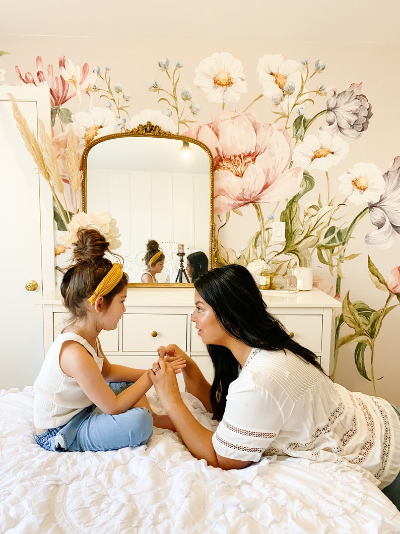 Puede incluir: Una madre y su hija est&aacute;n sentadas en una cama en una habitaci&oacute;n con un mural de pared floral. La hija lleva un top blanco sin mangas, pantalones vaqueros azules y una diadema amarilla. La madre lleva una blusa blanca con encaje y tiene el pelo largo y negro. Se est&aacute;n tomando de la mano y se miran.