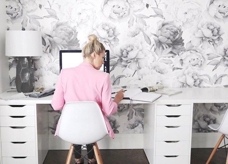 May include: A woman sits at a white desk with drawers, working on a notebook. The desk is in front of a wall with a floral wallpaper featuring large gray peonies.