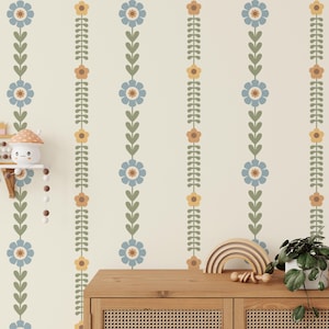 Puede incluir: Una pared de color crema con un patrón repetitivo de rayas florales verticales. El diseño presenta flores azules, amarillas y marrones, alternando con patrones de hojas y tallos verdes. Un mueble de madera con frente tejido está en primer plano.