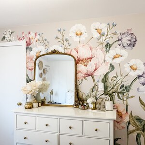 Puede incluir: Una c&oacute;moda blanca con seis cajones y pomos dorados se encuentra frente a una pared con un mural de papel tapiz floral. El mural presenta grandes flores rosas, blancas y moradas con hojas verdes. Un espejo dorado enmarcado con un borde biselado se encuentra sobre la c&oacute;moda.