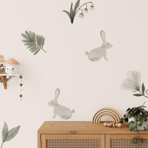 May include: Watercolor-style wall decals featuring two gray rabbits, leaves, and flowers. A wooden rainbow and a small plant in a white pot sit on a wooden cabinet. The background is a light beige color.