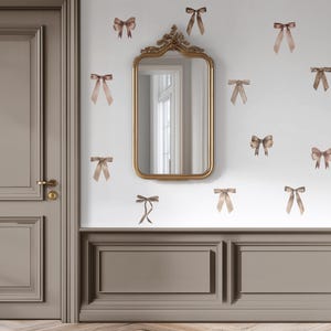 May include: A gold-framed mirror hangs on a white wall decorated with watercolour-style ribbon decals in shades of brown and tan. The wall also features decorative wainscoting. A closed door is visible on the left.
