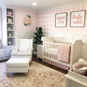 May include: A nursery room featuring a white crib with a pink quilted blanket, a white wingback chair with an ottoman, a white nightstand, and a rug with a red floral pattern. The walls have white wainscoting and pink and white gingham wallpaper.