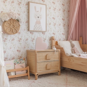 May include: A light pink heart-shaped nightlight sits on a woven rattan bedside table with two drawers. The bedside table is next to a wooden bed with a white and pink bedding set. The wall behind the bed is covered in a floral wallpaper with pink flowers.