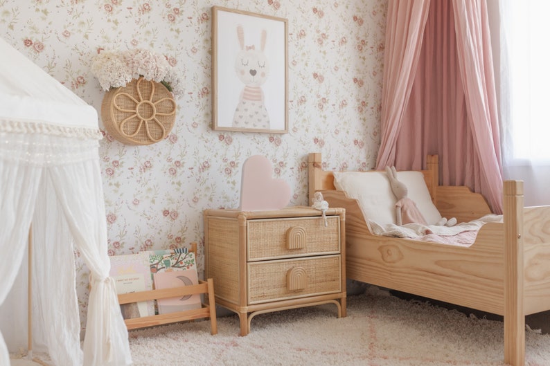 May include: A light pink heart-shaped nightlight sits on a woven rattan bedside table with two drawers. The bedside table is next to a wooden bed with a white and pink bedding set. The wall behind the bed is covered in a floral wallpaper with pink flowers.
