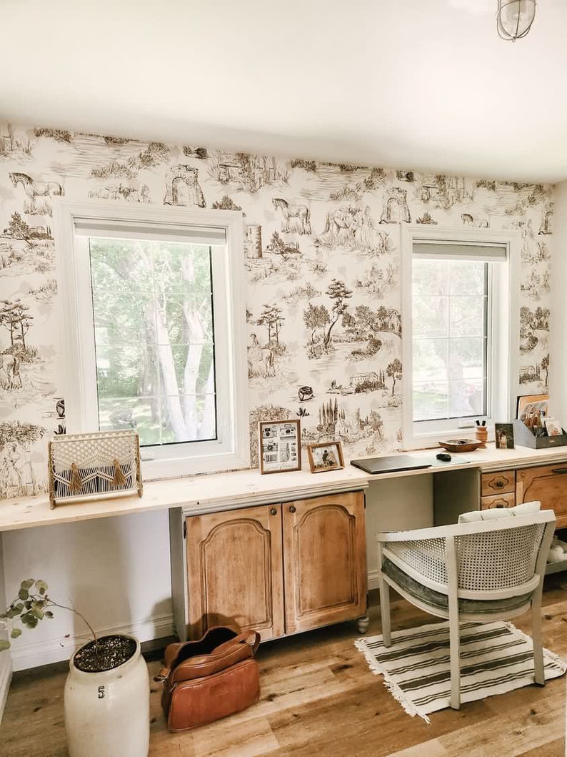 K&ouml;nnte beinhalten: Ein Heimb&uuml;ro mit einem hellen Holzschreibtisch und zwei St&uuml;hlen. Die W&auml;nde sind mit einer braunen und wei&szlig;en Tapete mit Toile-Muster bedeckt. Es gibt zwei Fenster mit wei&szlig;en Jalousien. Eine braune Ledertasche liegt auf dem Boden neben einer Topfpflanze.