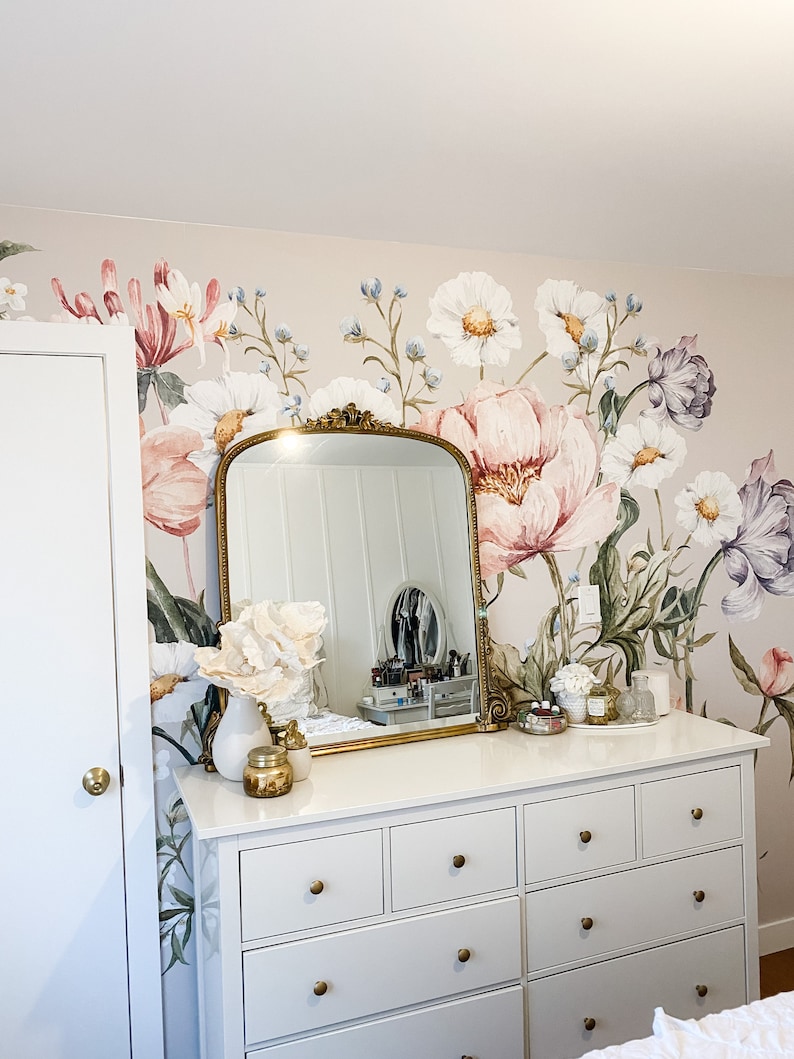 Puede incluir: Una c&oacute;moda blanca con pomos dorados y seis cajones. La c&oacute;moda est&aacute; decorada con un arreglo floral blanco, un candelero dorado y un espejo dorado. La c&oacute;moda est&aacute; frente a una pared con un papel tapiz floral con flores rosas, blancas y moradas.