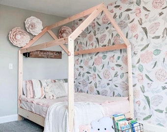 May include: A wooden bed frame in the shape of a house with a white blanket and pink and white pillows. The wall behind the bed is covered in floral wallpaper with pink and green flowers. The text "God is within her, she shall not fall" is written on a wooden sign above the bed.
