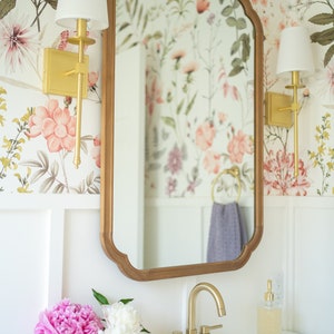 May include: A white bathroom vanity with a gold faucet and a mirror with a floral wallpaper background. Two gold sconces with white shades are on either side of the mirror. Two pink and white peonies are in glass vases on the counter.