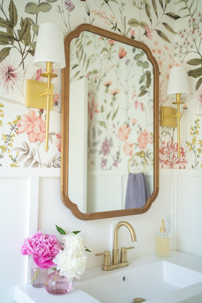 May include: A white bathroom vanity with a gold faucet and a mirror with a floral wallpaper background. Two gold sconces with white shades are on either side of the mirror. Two pink and white peonies are in glass vases on the counter.