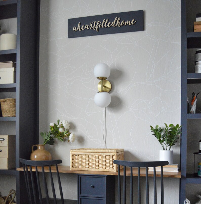 May include: A home office desk with a black and wood finish. The desk has a wicker storage box, a gold wall sconce with two white globe lights, and a potted plant. The wall behind the desk has a white and gray abstract wallpaper with the text "aheartfilledhome" on a black sign.