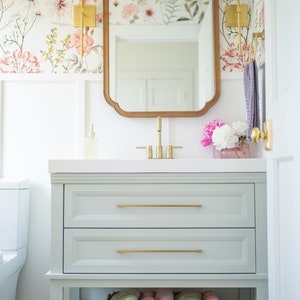 May include: A light green bathroom vanity with two drawers and a lower shelf. The vanity has gold hardware and a white countertop. A mirror with a wood frame hangs above the vanity. The wall behind the vanity is covered in floral wallpaper.